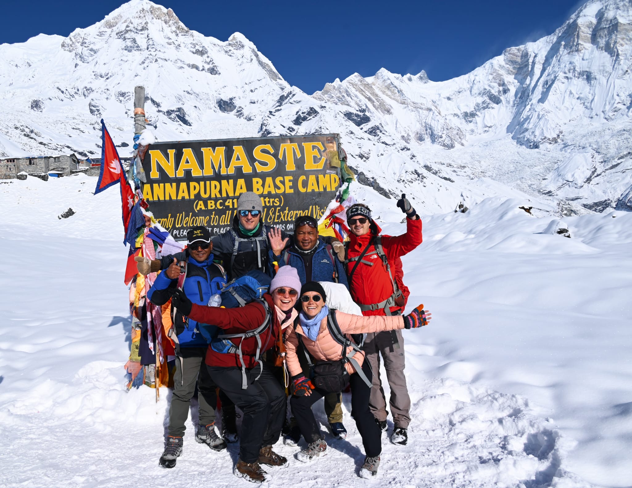 Trekkers celebrating at Annapurna Base Camp sign surrounded by snow and Himalayan peaks.