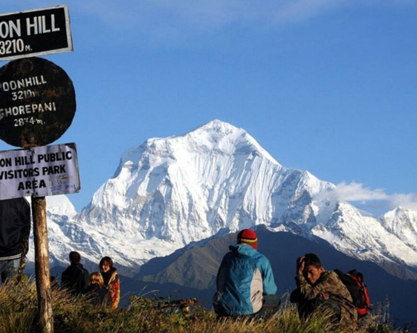 Trekkers sitting at Poon Hill viewpoint looking toward snow-capped Dhaulagiri mountain peak in the Himalayas, Nepal, with directional signs nearby.