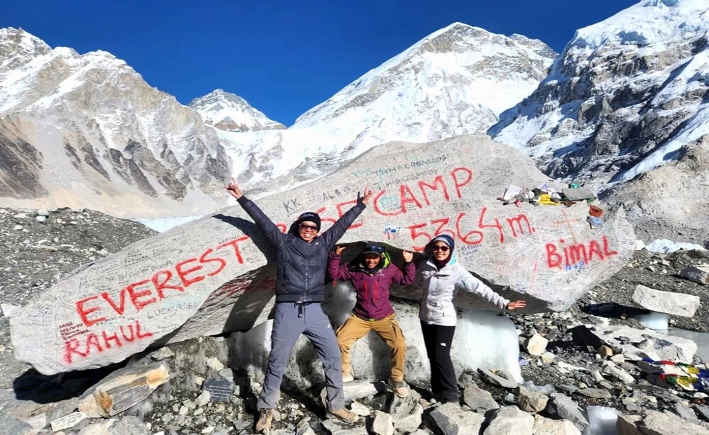 Three trekkers celebrate at Everest Base Camp marker stone with the Himalayas and snow-covered peaks in the background.