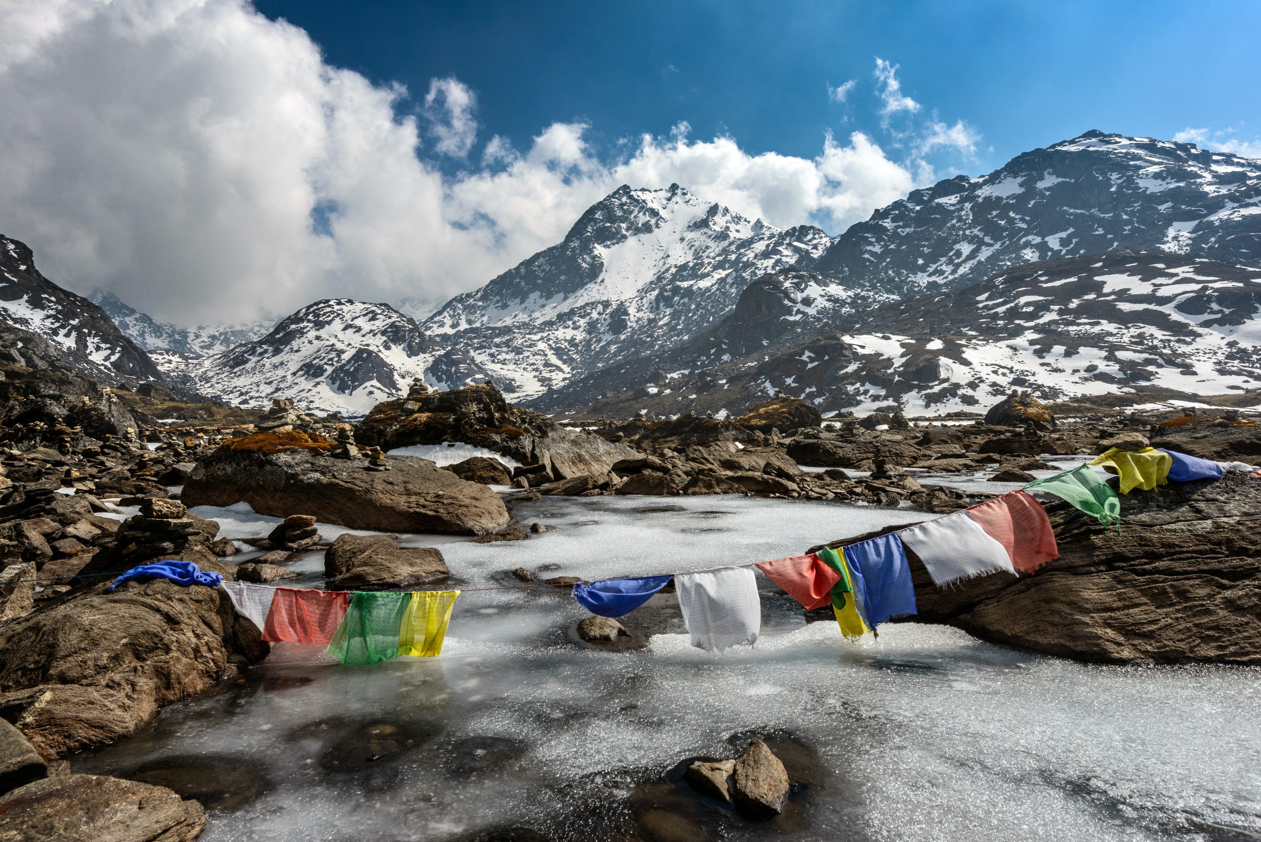 Snow-covered mountain landscape in Nepal with colorful prayer flags hanging off rocks with a partially frozen stream in the foreground.